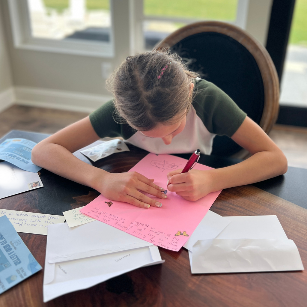 Child writing on a pink sheet of paper at a table with envelopes and papers around.