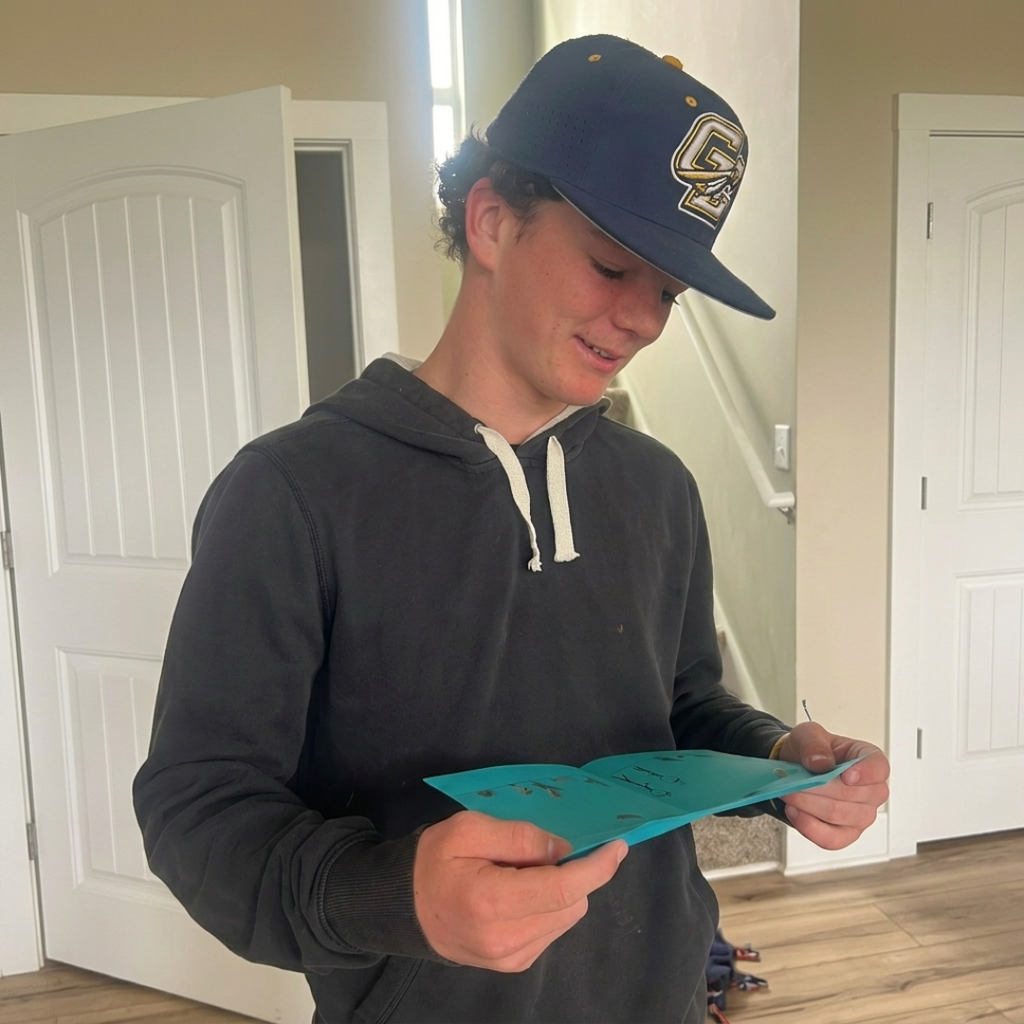 Person wearing a navy cap and gray hoodie holding a blue piece of paper indoors- reading a letter.